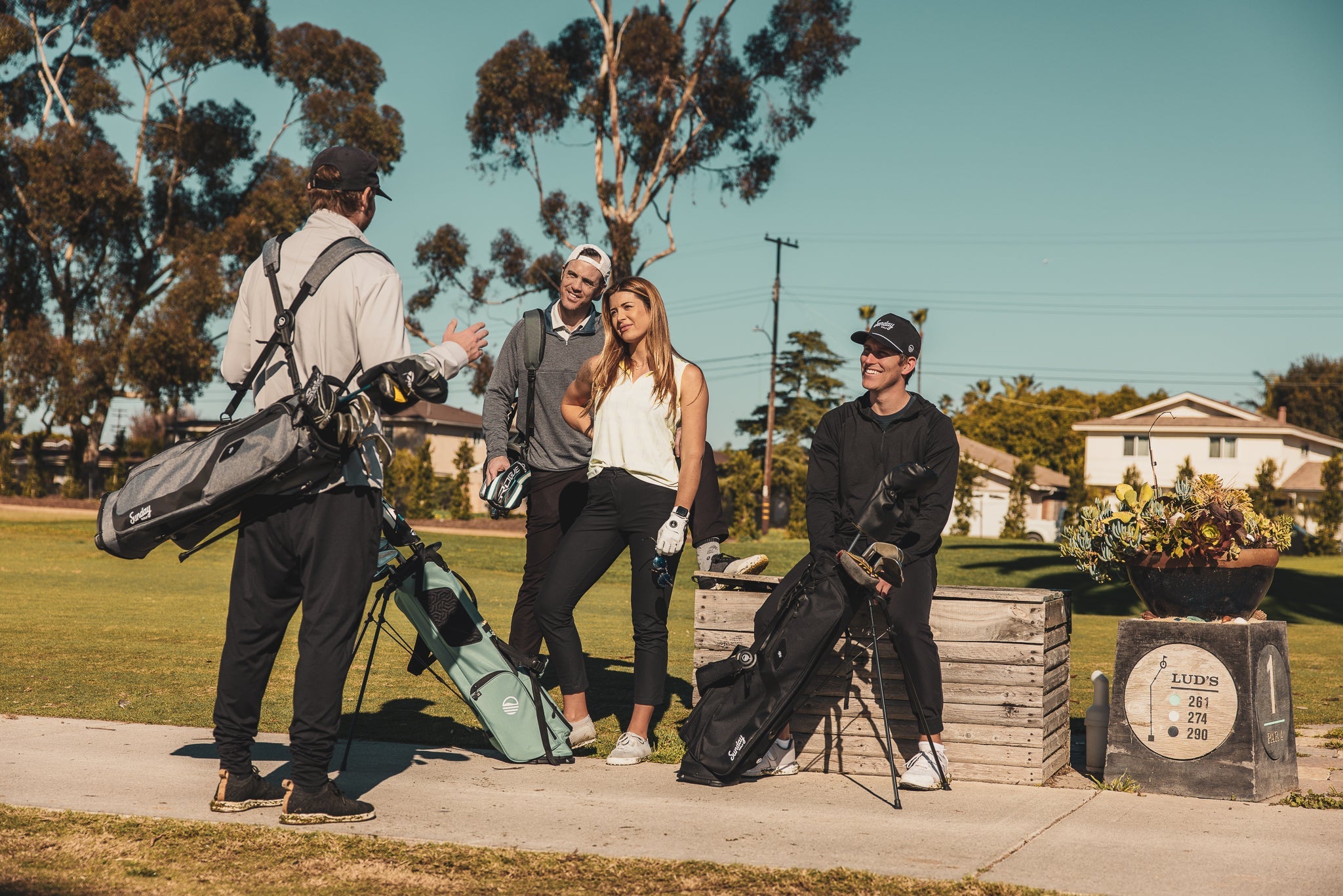 man standing with heather gray El Camino golf bag by Sunday Golf, woman standing with seafoam El Camino golf bag by Sunday Golf, and man sitting with  matte black El Camino golf bag by Sunday Golf